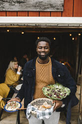 Portrait of smiling man with food standing at doorway during social gathering - MASF21204