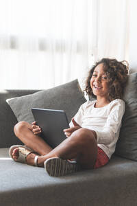 Porträt eines kleinen Mädchens lächelnd auf dem Sofa mit digitalem Tablet in den Händen - JAQF00113