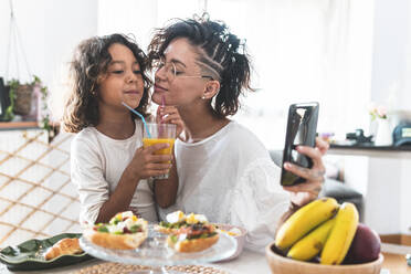 Mother and daughter taking selfie during breakfast - JAQF00122