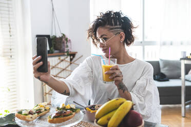 Porträt einer jungen Frau, die ein Selfie mit ihrem Smartphone macht, während sie frühstückt - JAQF00129