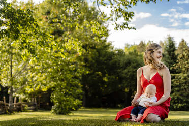 Smiling mother holding baby boy while kneeling on grassy land in park - AWAF00024