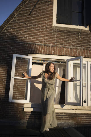 Cheerful woman with arms outstretched screaming while standing by window outside house stock photo