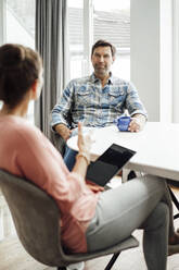 Smiling mature man looking at woman while talking on dining table in home - JOSEF02705