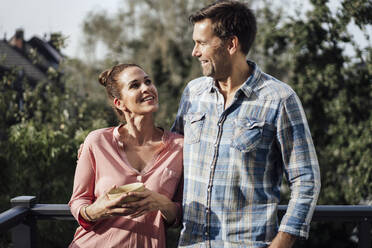 Happy mature couple looking at each other while standing in balcony on sunny day - JOSEF02706