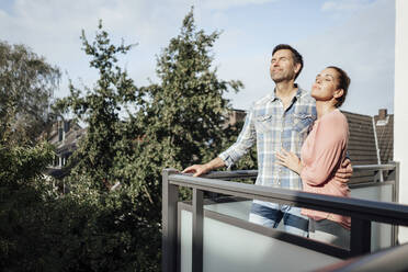 Mature couple with eyes closed standing in balcony together on sunny day - JOSEF02722