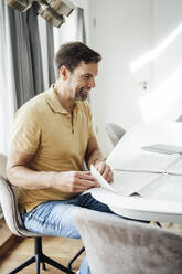Happy mature man turning page of newspaper on table at home - JOSEF02734
