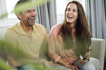 Fröhliches reifes Paar, das lachend auf dem Sofa in der Wohnung sitzt - JOSEF02737