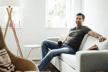 Smiling mature man holding coffee cup while sitting on sofa at home - JOSEF02743