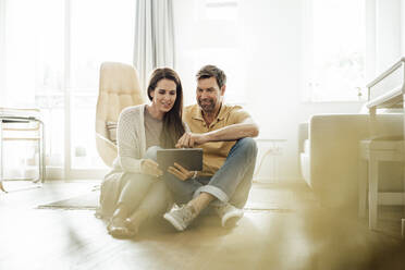 Smiling mature couple using digital tablet together while sitting on floor at home - JOSEF02756