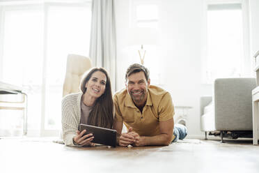 Smiling mature man and woman with digital tablet lying on floor in apartment - JOSEF02757