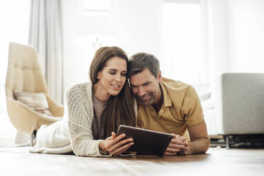 Lächelndes reifes Paar, das gemeinsam ein digitales Tablet benutzt, während es auf der Vorderseite einer Wohnung liegt - JOSEF02758