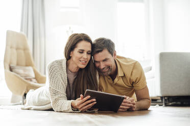 Smiling mature couple using digital tablet together while lying on front in apartment - JOSEF02758