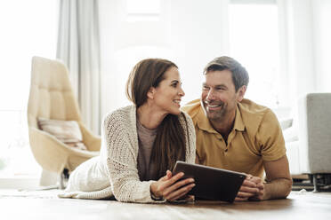 Smiling man and woman with digital tablet looking at each other while lying in home - JOSEF02759