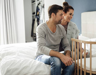 Smiling mature couple admiring crib while sitting on bed at home - JOSEF02760
