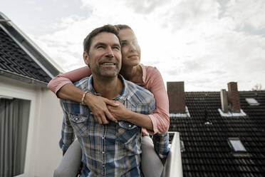 Smiling mature man carrying woman piggyback in balcony against cloudy sky - JOSEF02773