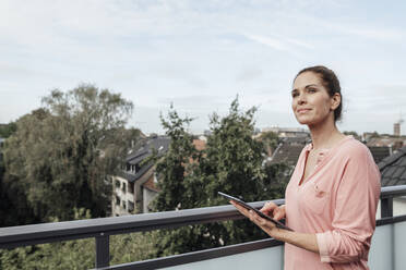 Lächelnde reife Frau hält digitales Tablet, während sie auf dem Balkon träumt - JOSEF02775