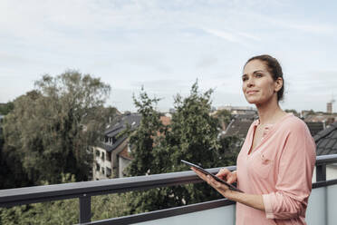 Smiling mature woman holding digital tablet while day dreaming in balcony - JOSEF02775