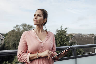 Mature woman contemplating while holding digital tablet against sky in balcony - JOSEF02776