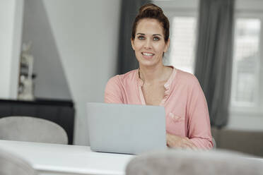 Happy female entrepreneur with laptop at table in home office - JOSEF02781