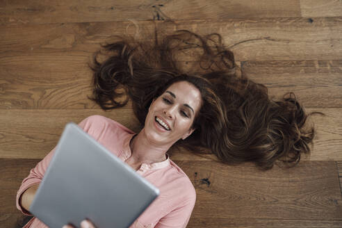 Glückliche reife Frau, die ein digitales Tablet benutzt, während sie in der Wohnung auf dem Rücken liegt - JOSEF02782