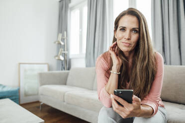 Mature woman with hand in hair holding mobile phone in living room at home - JOSEF02790