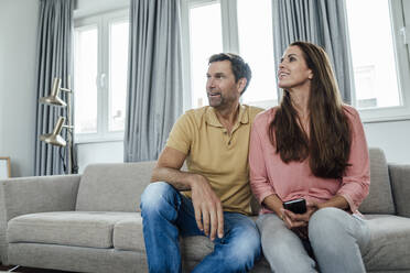 Smiling mature couple looking away while sitting on sofa in apartment - JOSEF02793