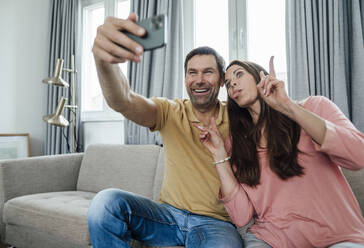 Happy mature man taking selfie while woman making funny face at home - JOSEF02795