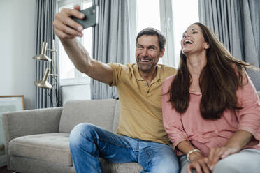 Älteres Paar lacht und nimmt Selfie beim Sitzen auf Sofa im Wohnzimmer - JOSEF02796