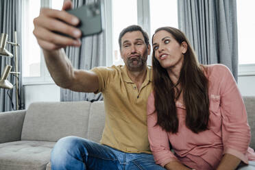 Glückliches Paar macht Gesichter bei der Aufnahme eines Selfies, während es auf dem Sofa zu Hause sitzt - JOSEF02797