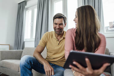 Reife Frau mit digitalem Tablet, die einen glücklichen Mann ansieht, während sie auf dem Sofa in einer Wohnung sitzt - JOSEF02802