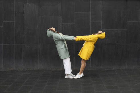 Sisters wearing raincoats holding hands while standing against black wall - AXHF00014