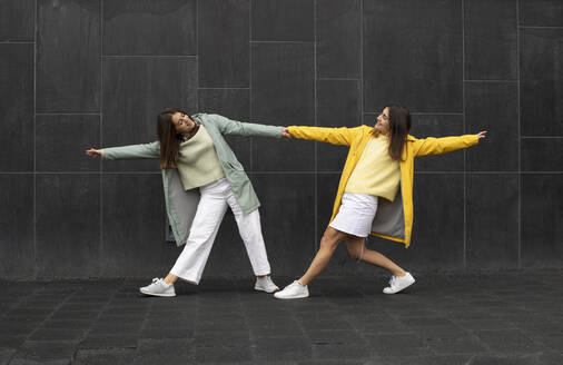 Sisters wearing blue and yellow raincoats holding hands while dancing on footpath - AXHF00015