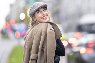 Glückliche Frau, die einen Mantel hält und über die Schulter schaut, während sie auf der Straße steht - GGGF00706