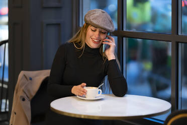 Lächelnde Frau mit Kaffeetasse im Gespräch auf dem Smartphone in einem Café bei Nacht - GGGF00728