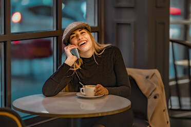 Frau lacht mit Kaffeetasse, während sie in einem Café mit ihrem Smartphone telefoniert - GGGF00729