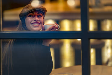 Lächelnde Frau hält eine Kaffeetasse und schaut durch das Fenster eines Cafés - GGGF00731