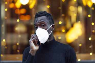 Man wearing protective face mask talking on mobile phone while standing outdoors during Covid-19 - PGF00336