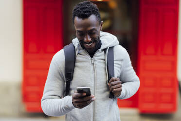 Young man with backpack smiling while using mobile phone standing outdoors - PGF00345