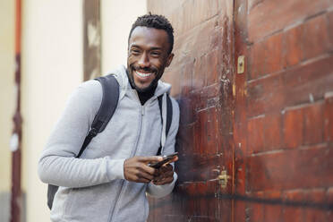 Young man with backpack and mobile phone smiling while leaning on wall - PGF00348