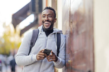 Smiling man using mobile phone while leaning on wall - PGF00349