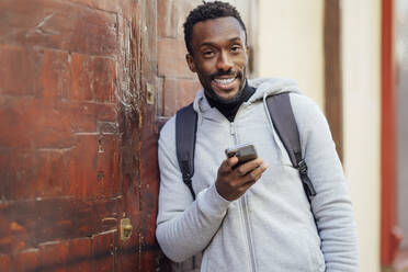 Man with backpack and mobile phone smiling while leaning on wall - PGF00350