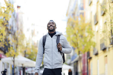 Young man with backpack smiling while walking in city - PGF00364