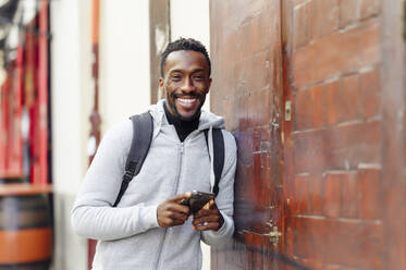 Man with backpack and mobile phone smiling while leaning on wall - PGF00366