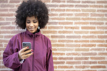 Smiling young woman using smart phone while standing against brick wall - IFRF00242