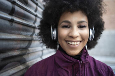 Glückliche junge Frau hört Musik über Kopfhörer an einer Wellblechwand - IFRF00254