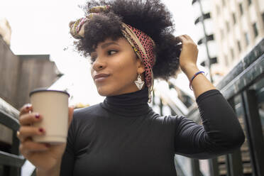 Junge Afro-Frau mit Hand im Haar, die wegschaut, während sie eine Einweg-Kaffeetasse in der Stadt hält - IFRF00270