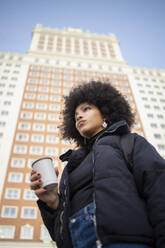 Afro junge Frau nachdenklich, während hält Kaffeetasse gegen gebaut Struktur in der Stadt - IFRF00276