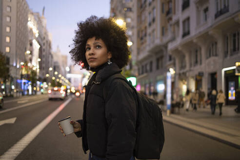 Young woman with disposable coffee cup crossing road while looking around in city at night - IFRF00282