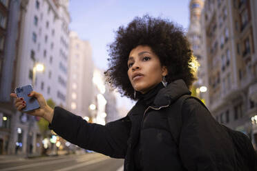 Afro-Frau mit Handy in der Hand, während sie nachts auf einer Straße in der Stadt eine Mitfahrgelegenheit sucht - IFRF00283