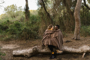 Couple wrapped in blanket sitting face to face on fallen tree at forest - EGAF01379